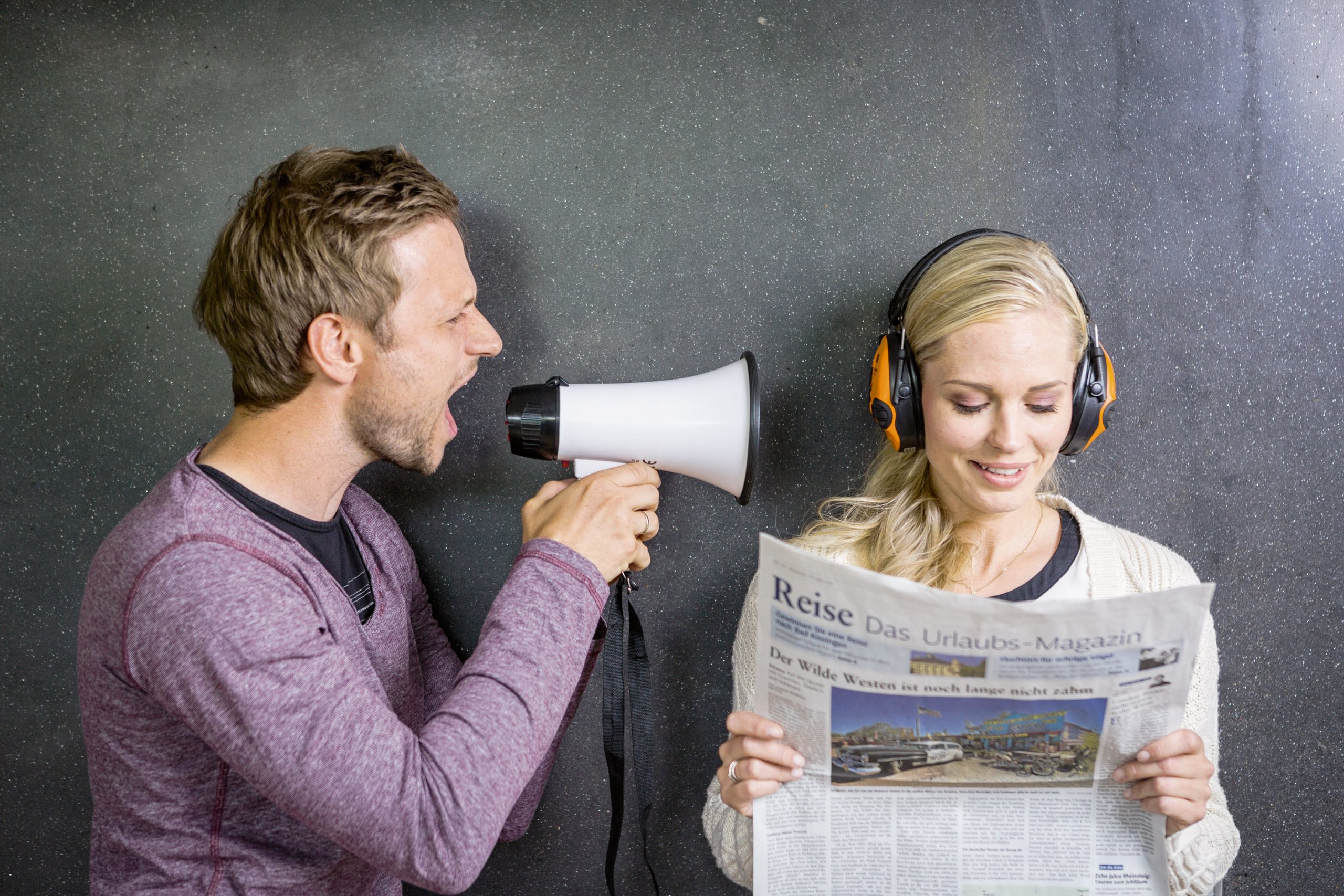 Mann schreit mit einem Megaphon eine Frau an, die Kopfhörer trägt und eine Zeitung liest.